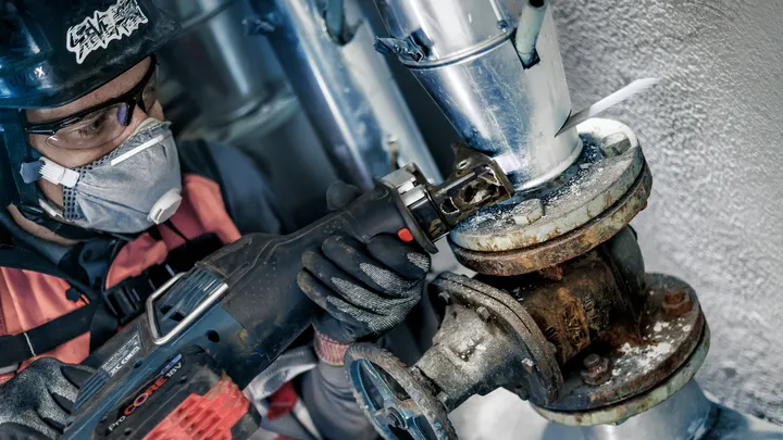 A person wearing safety equipment uses a reciprocating saw to cut a corroded pipe.