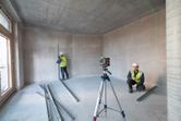Two people wearing safety equipment use a laser leveling tool in an unfinished room.