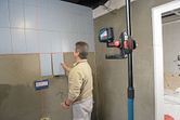 Person aligns wall tiles using a laser leveling tool mounted on a telescopic pole.