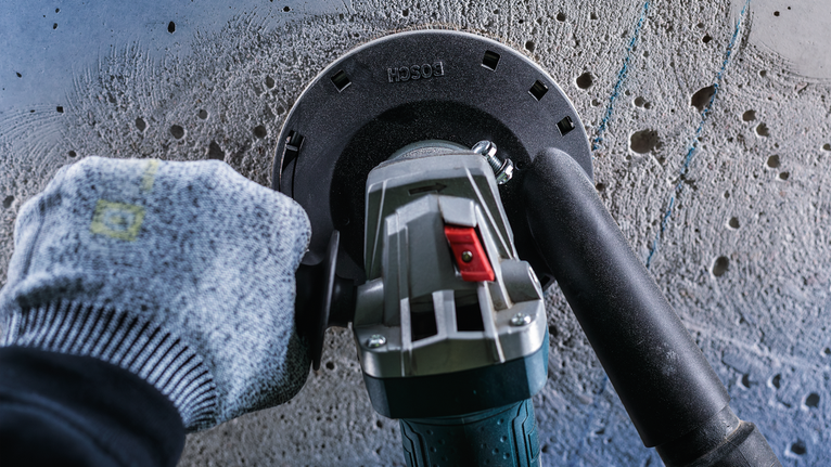 A worker wearing safety equipment grinds a concrete wall with a power tool.
