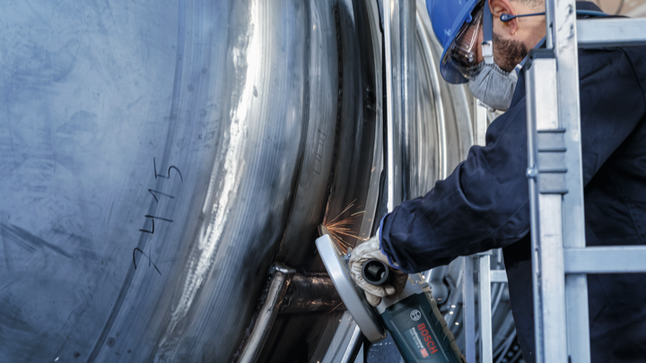 A person wearing safety equipment grinds a large metal tank, creating sparks.