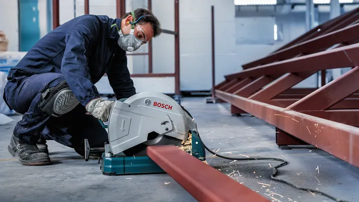 A person wearing safety equipment cuts a metal beam with a power saw, sparks flying.