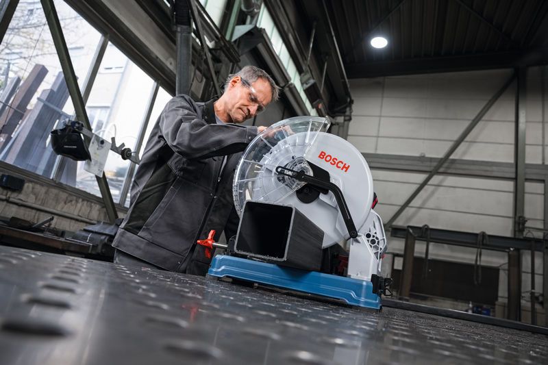 A person wearing safety equipment prepares a metal cut-off saw to cut a metal pipe.