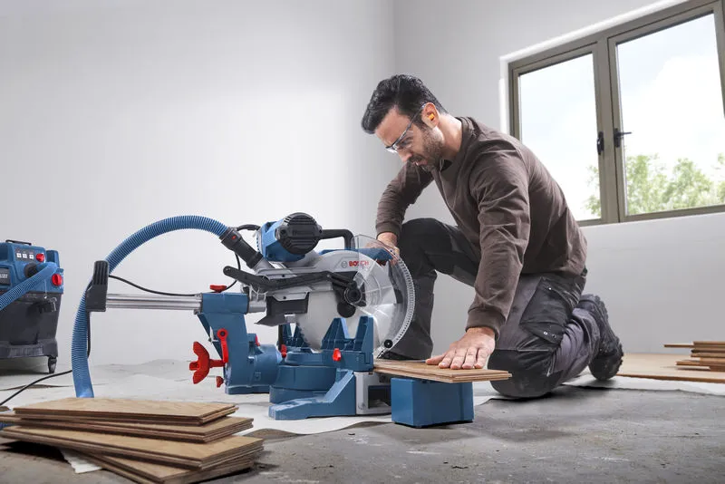 A person wearing safety equipment cuts wooden flooring with a mitre saw in a bright room.