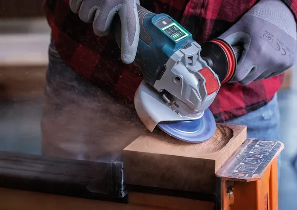 Worker wearing safety equipment uses an angle grinder to carve a circular hole in wood.