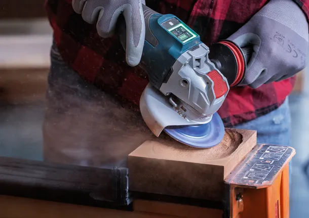 Worker wearing safety equipment uses an angle grinder to carve a circular hole in wood.
