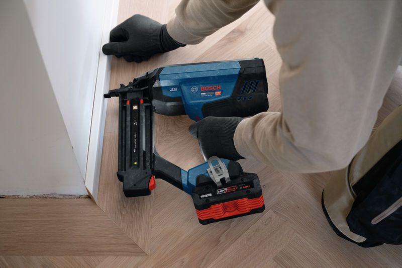 A person wearing safety equipment uses a cordless nailer to fasten floor trim.