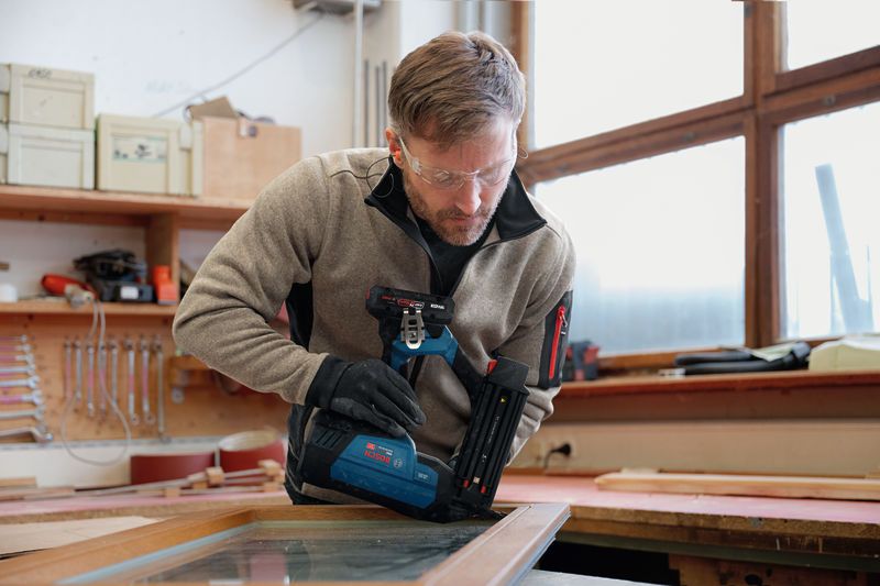 A worker wearing safety equipment uses a cordless nailer on a wooden window frame.