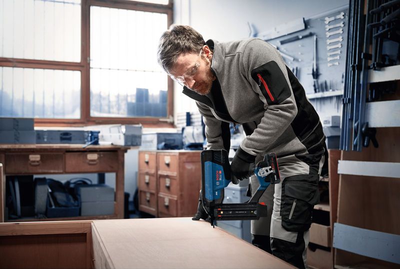A person wearing safety equipment uses a cordless nailer on wooden furniture.