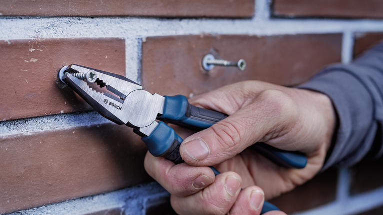Person uses pliers to grip a screw fastened into a brick wall.