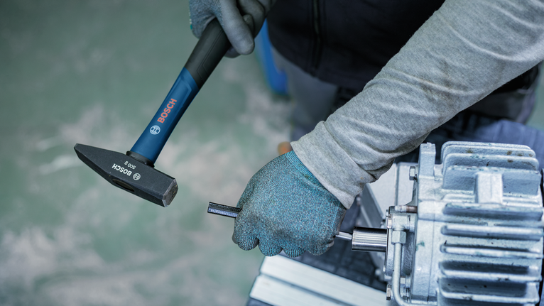Person wearing safety equipment uses a hammer on a metal shaft near machinery.