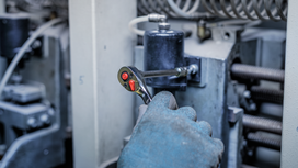 Person wearing safety equipment tightening a bolt with a ratchet wrench on industrial machinery.