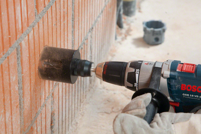 Worker wearing safety equipment drills a circular hole in a brick wall.