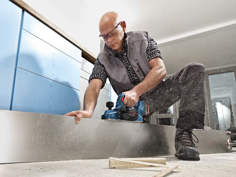 Una persona que usa equipo de seguridad cepilla una tabla de madera utilizando una cepilladora inalámbrica.