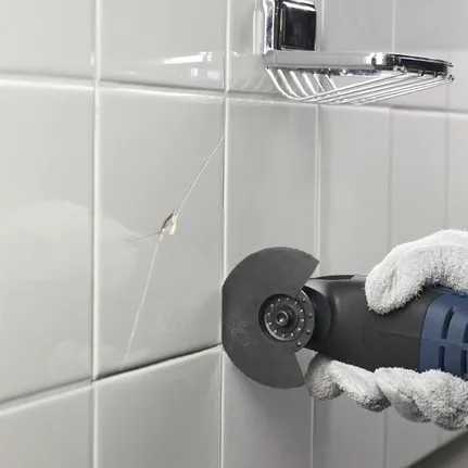 Una persona que lleva equipo de seguridad utiliza una herramienta oscilante para cortar azulejos agrietados del baño.