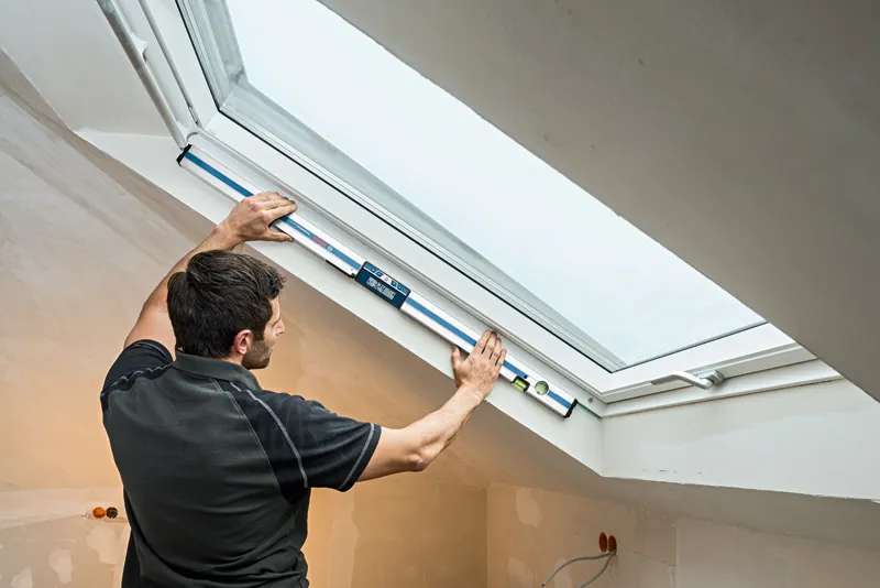 Persona midiendo el ángulo de una ventana con un inclinómetro digital en un ático terminado.
