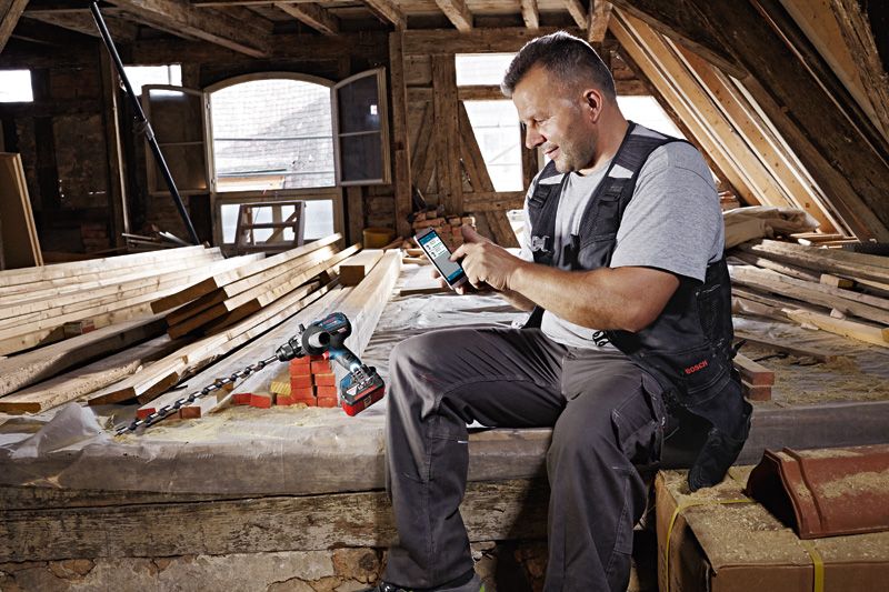 Una persona que usa equipo de seguridad usa un teléfono inteligente junto a un taladro inalámbrico en un taller de madera.