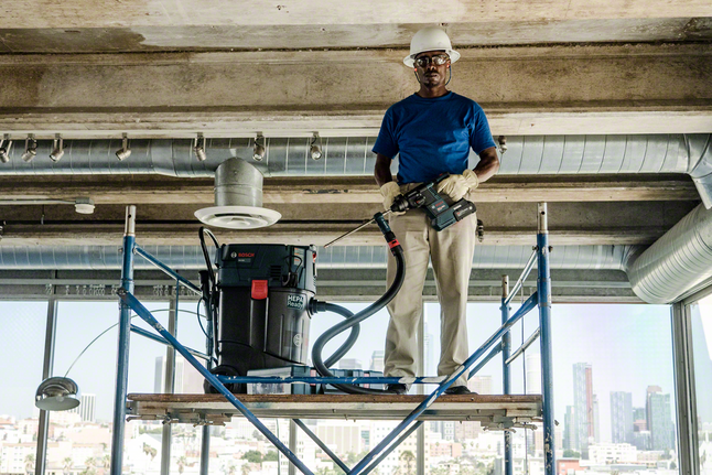 Una persona, que lleva puesto el equipo de seguridad, se encuentra sobre un andamio manejando una herramienta eléctrica con extractor de polvo.