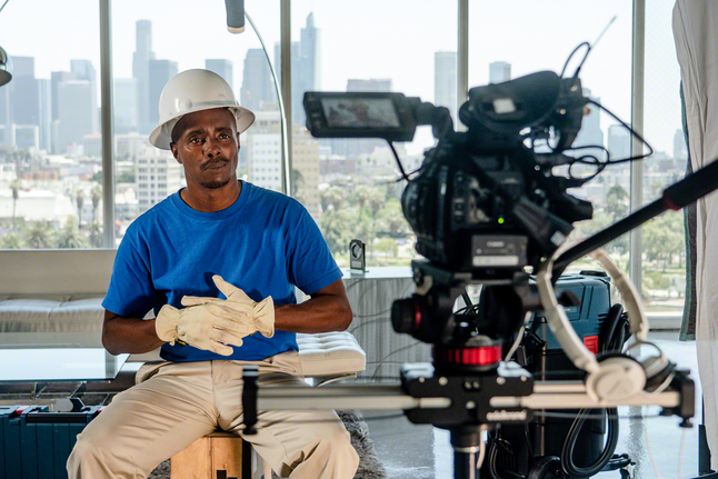 Una persona que lleva equipo de seguridad está sentada frente a una cámara de vídeo con edificios de la ciudad al fondo.