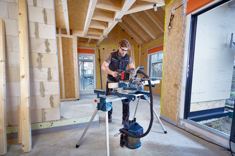 Una persona que usa equipo de seguridad corta madera con una sierra ingletadora inalámbrica en una casa en construcción.