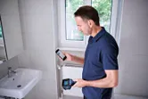 Persona que usa una cámara térmica y un teléfono inteligente en un baño alicatado.