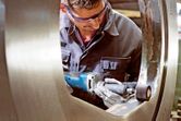 Una persona que usa equipo de seguridad pule un anillo grande de metal con una amoladora recta inalámbrica.