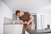 Una persona que usa equipo de seguridad utiliza un taladro combinado inalámbrico en un armario de cocina.
