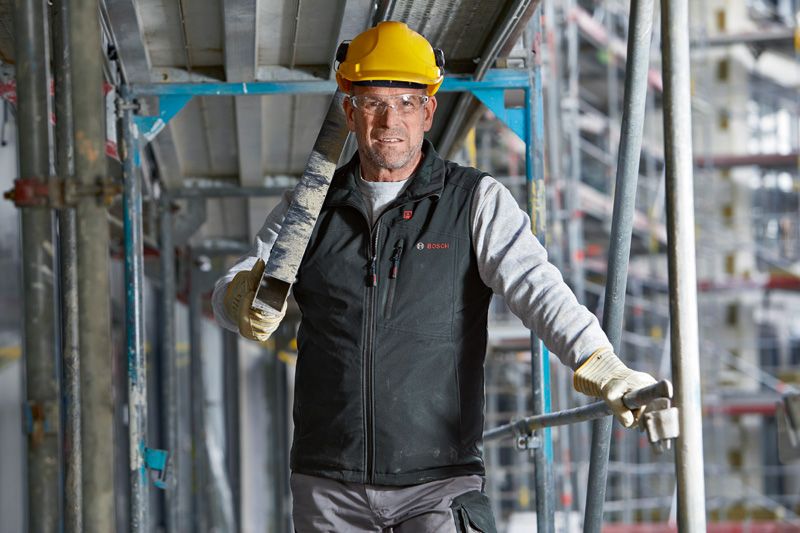 Una persona que usa equipo de seguridad y chaleco calefactado lleva una viga de metal en un sitio de construcción.