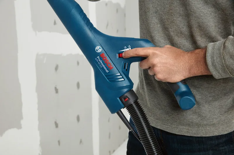 Una persona ajusta una herramienta eléctrica azul con una manguera cerca de un panel de yeso sin terminar.