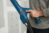 Una persona ajusta una herramienta eléctrica azul con una manguera cerca de un panel de yeso sin terminar.