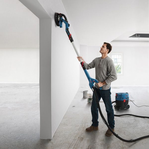 Una persona lija una pared blanca con una lijadora de yeso conectada a un extractor de polvo.