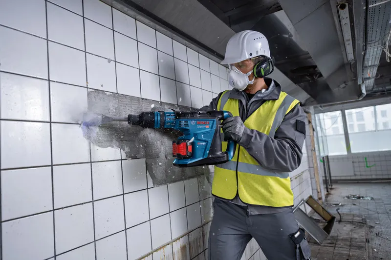 Una persona que usa equipo de seguridad utiliza un martillo rotatorio inalámbrico para romper azulejos de la pared.