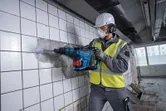 Una persona que usa equipo de seguridad utiliza un martillo rotatorio inalámbrico para romper azulejos de la pared.