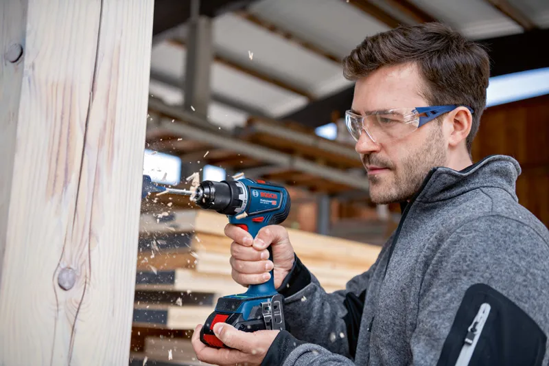 Una persona que usa equipo de seguridad perfora una viga de madera en un taller.