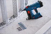 Pistola de clavos inalámbrica colocada en el piso al lado de tiras de fijación y montantes de estructura de metal.