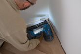 Una persona que usa equipo de seguridad utiliza una clavadora de madera inalámbrica para fijar la moldura al piso.