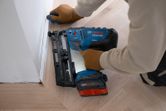 Una persona que usa equipo de seguridad usa una clavadora de madera inalámbrica para fijar la moldura al piso.