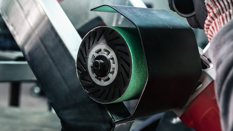 Una persona que usa equipo de seguridad opera una lijadora de banda sobre una superficie metálica.