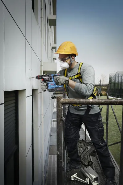 Una persona que usa equipo de seguridad perfora una pared exterior mientras está parada sobre un andamio.