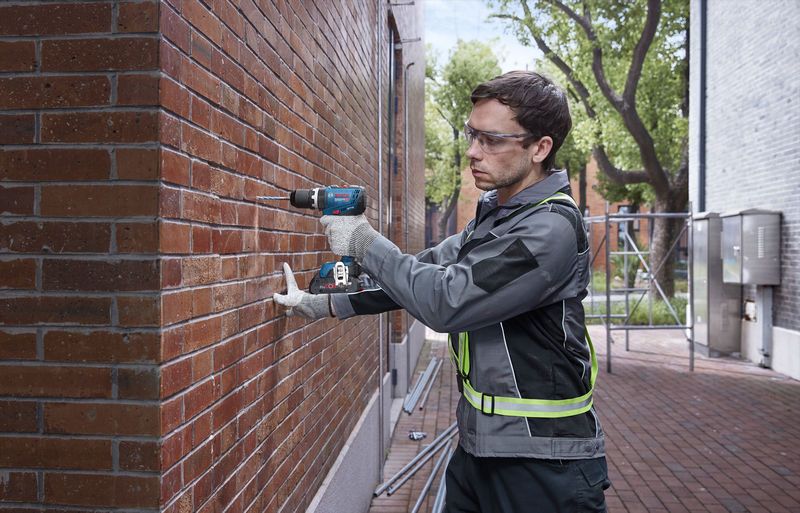 Una persona que usa equipo de seguridad perfora una pared de ladrillos al aire libre.