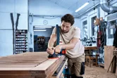 Una persona que usa equipo de seguridad lija una superficie de madera con una lijadora orbital aleatoria inalámbrica.