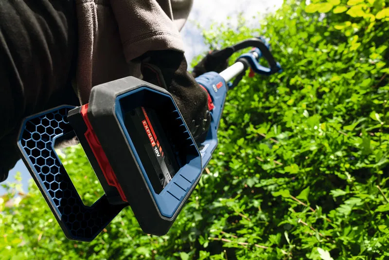 Una persona que usa equipo de seguridad corta arbustos verdes densos con un cortasetos inalámbrico.