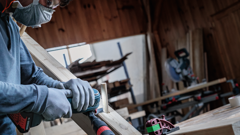 Una persona que usa equipo de seguridad lija una tabla de madera con una herramienta eléctrica.