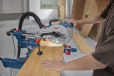 Una persona opera una sierra ingletadora para cortar una tabla de madera en un taller.
