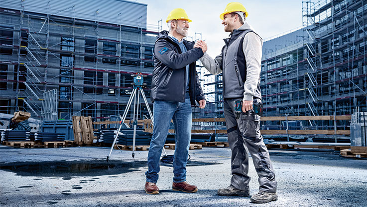 Dos hombres con cascos amarillos se saludan en un sitio de construcción.