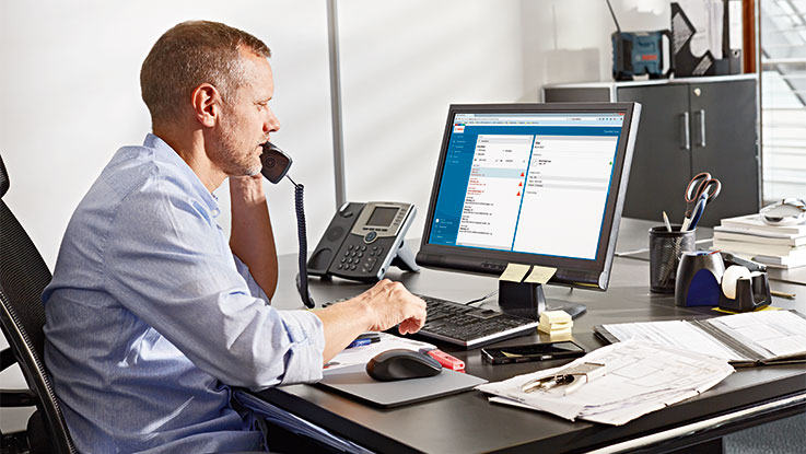 Hombre con camisa azul en escritorio, hablando por teléfono, usando computadora.