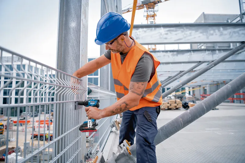 Turvavarusteita käyttävä henkilö kiinnittää metalliaidan akkukäyttöisellä iskuruuvinvääntimellä.