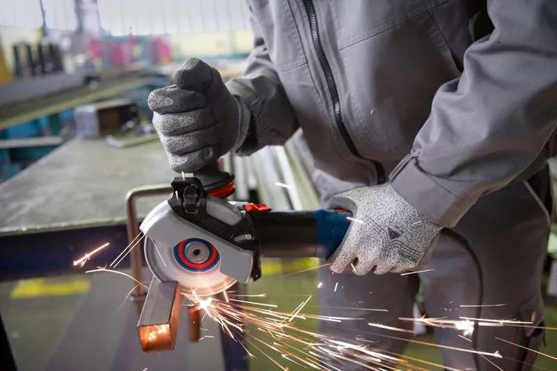 Turvavarusteita käyttävä henkilö hioo metallitankoa käsikulmahiomakoneella.