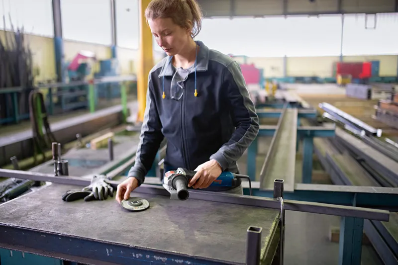 Turvavarusteita käyttävä henkilö valmistelee kulmahiomakonetta metallityöpöydällä.