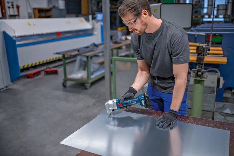 Turvavarusteita käyttävä henkilö leikkaa metallilevyä akkukäyttöisellä sähkötyökalulla.